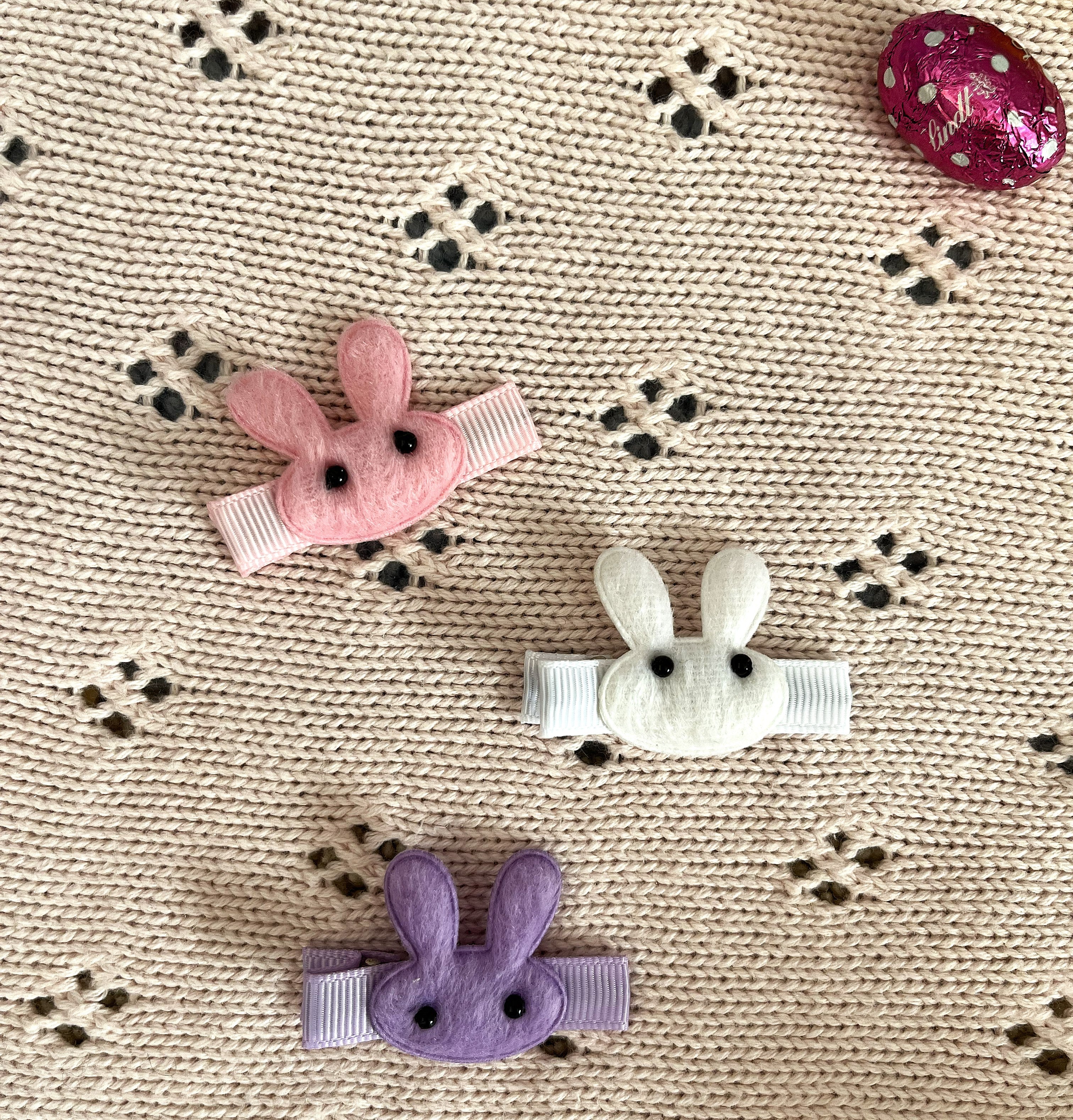 Three bunny-shaped hair clips in pink, white, and purple on a pink blanket 